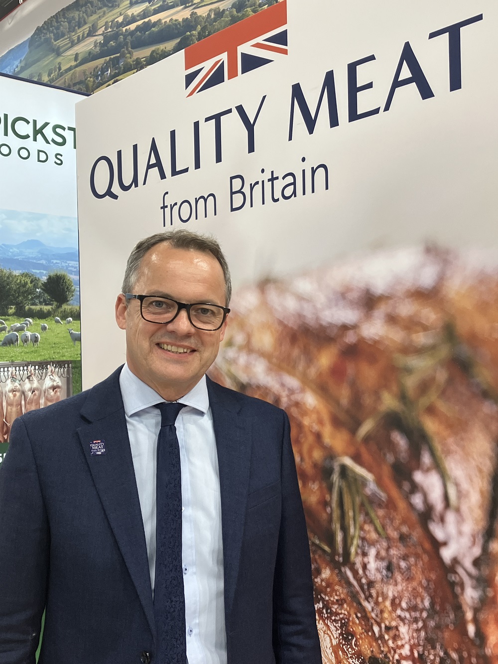 Man (Graham Wilkinson) with glasses wearing dark suit and tie stood in front of Quality Meat poster